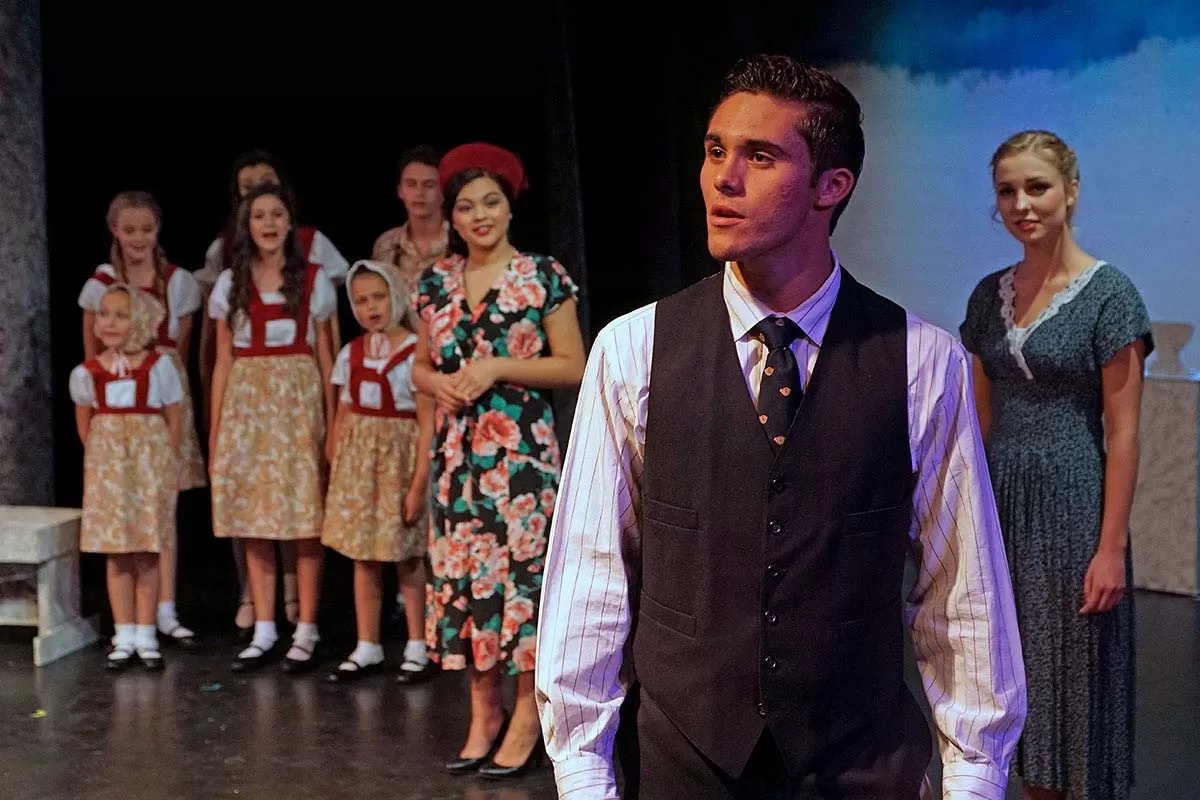 A stage performance with a man in a suit in the foreground, and people in costume, including children, in the background.