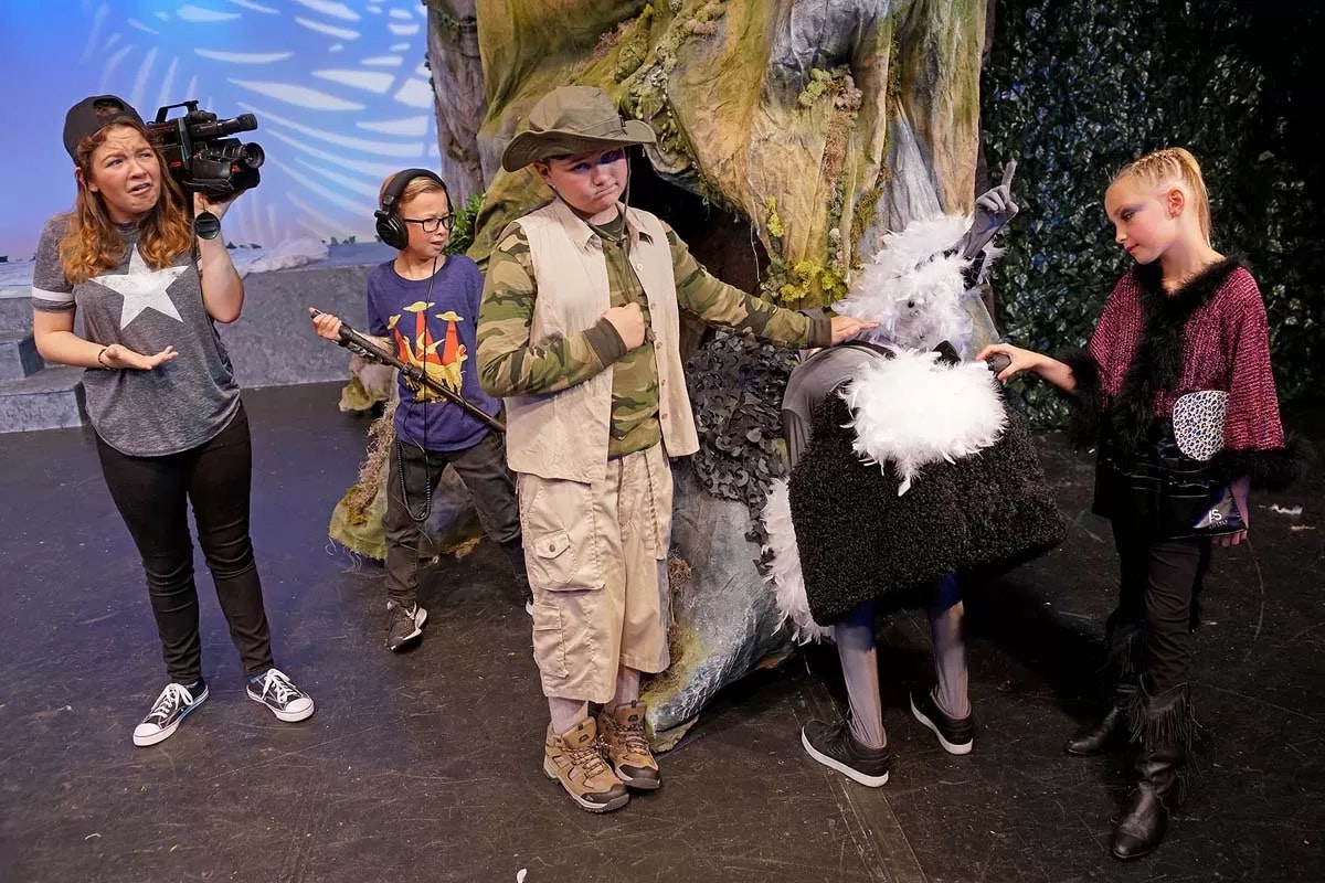 A group of kids in costumes on a stage with a person filming them. One is dressed as a ram, others in various roles, mid-performance.