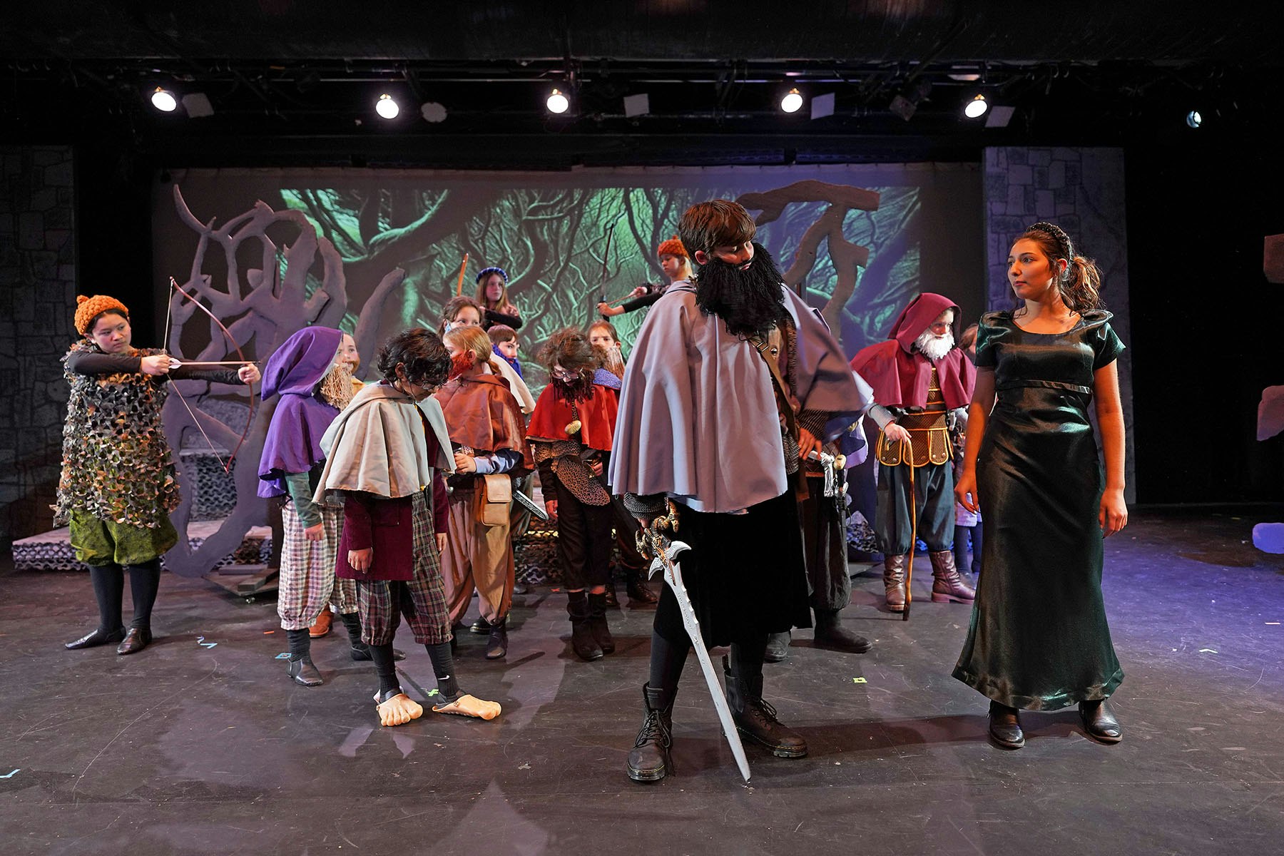 A group of people in medieval costumes, holding props like a bow and sword, stand on a stage with a forest backdrop.