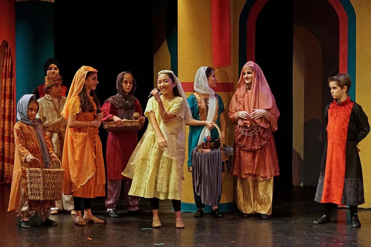 A group of people in colorful, traditional costumes on stage, possibly performing a theatrical play.