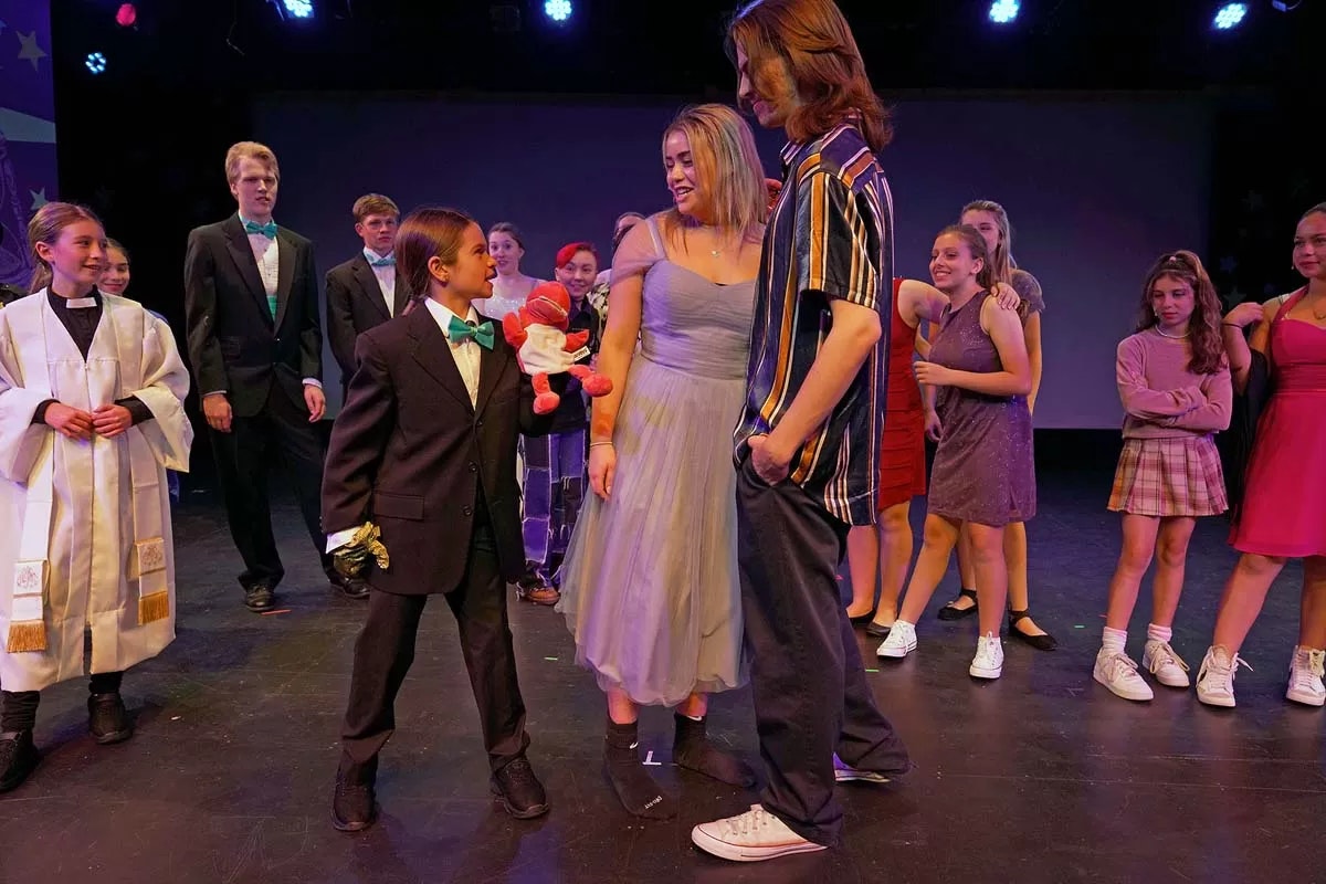 A group of people on stage in various costumes, including formal and casual attire, with colorful lighting overhead.