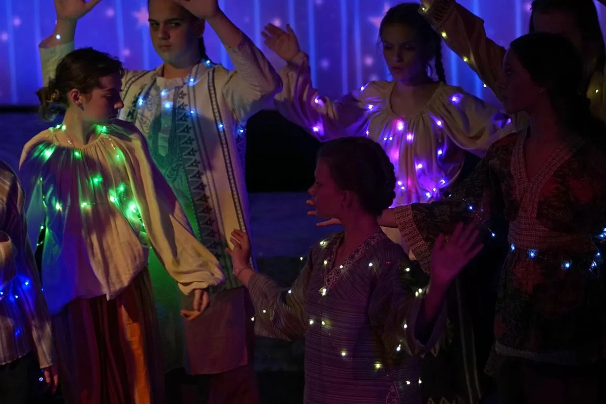 People in traditional attire adorned with colorful LED lights, standing with hands raised in a dimly lit setting.
