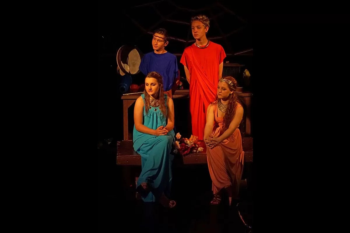 Four people in colorful robes and headbands are posed in a theatrical setting with drums in the background.