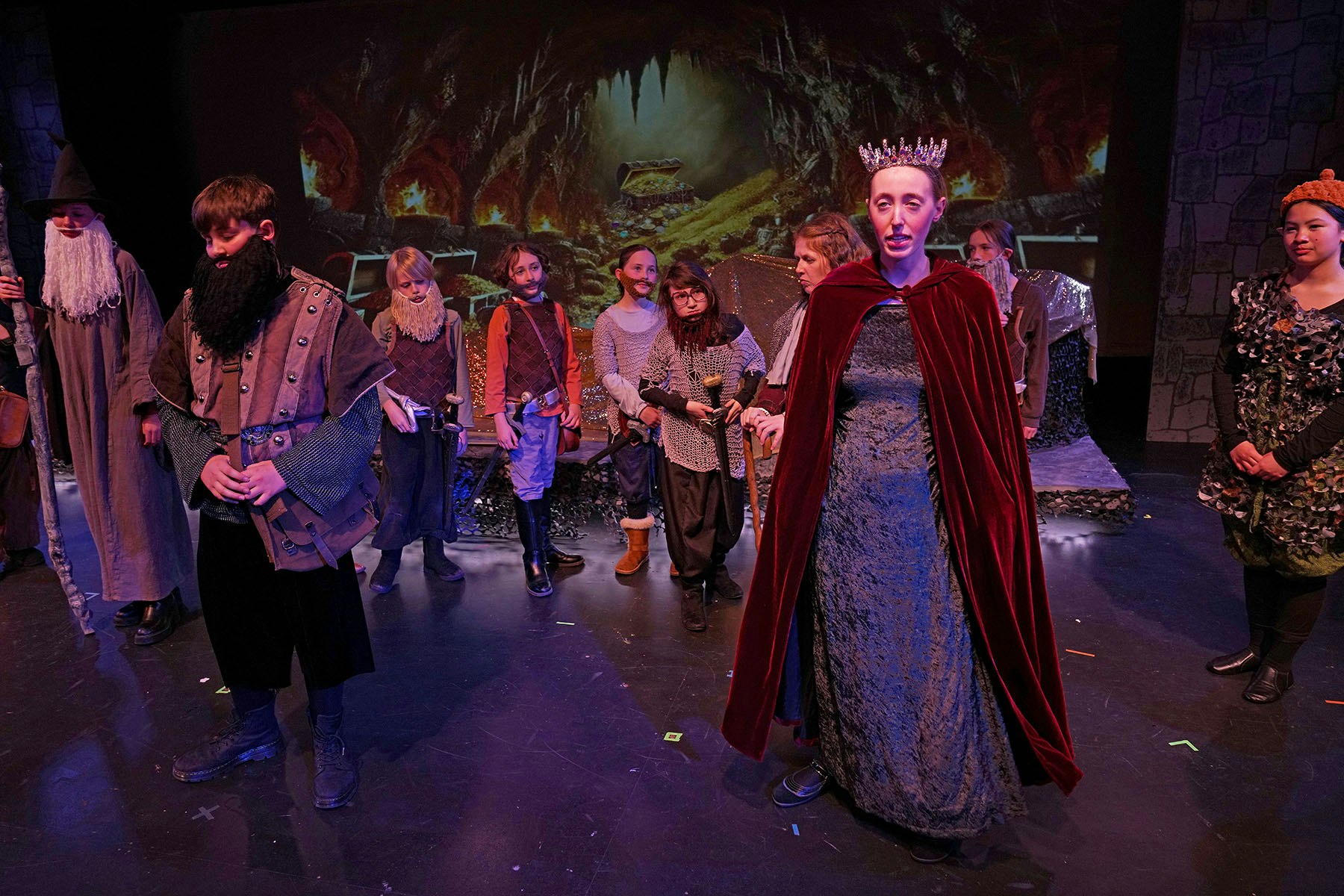 Group of people in fantasy costumes on a stage with a cave backdrop, including a person in a crown and beard.