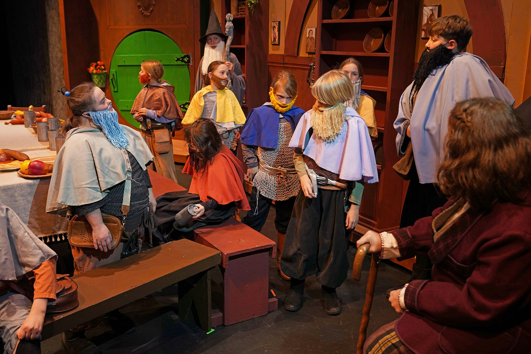 A group of children in costumes, bearded and caped, are gathered in a set with a green door and medieval decor, resembling a play scene.