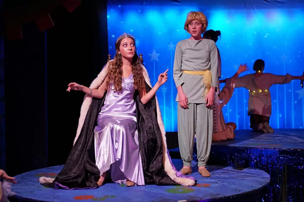 Two people on stage; one in a purple gown and tiara seated, the other in a light outfit standing. Starry background.