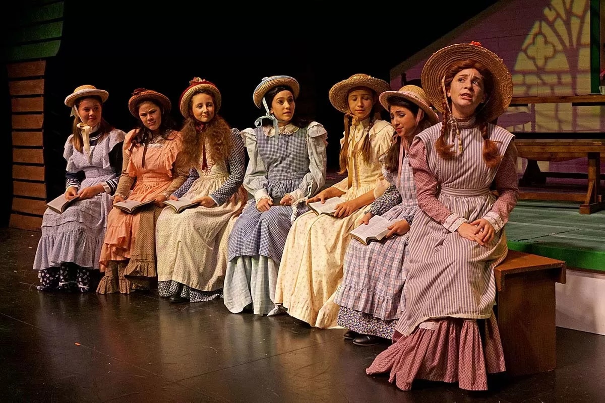 A group of seven women in vintage dresses and hats sit with open books on stage.