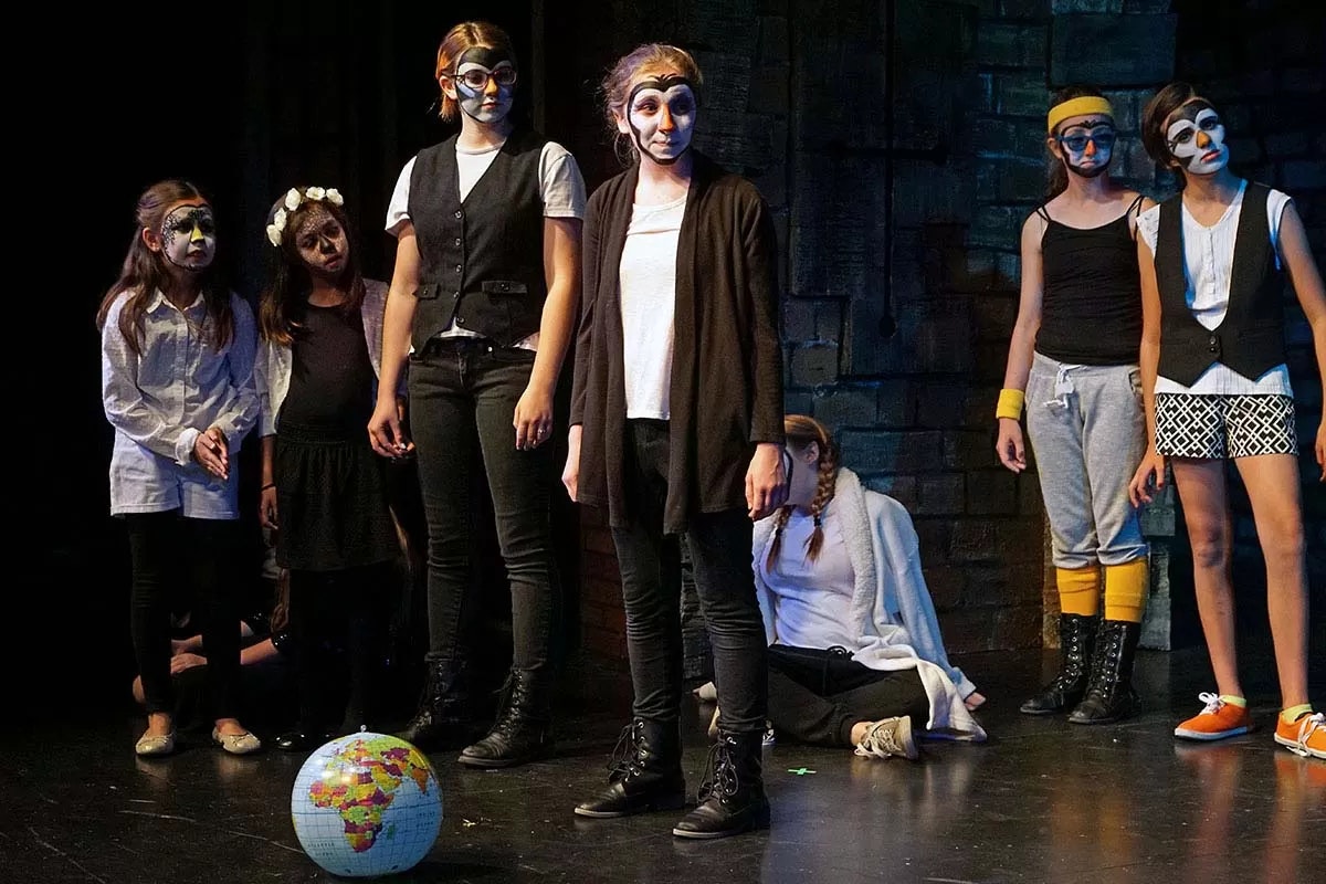 A group of children in theatrical costumes and face paint stand on stage with a globe at their feet.