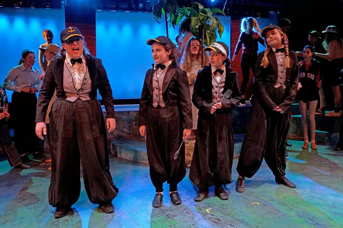 A group of performers in tuxedos and caps, possibly in a play, with a colorful stage background.