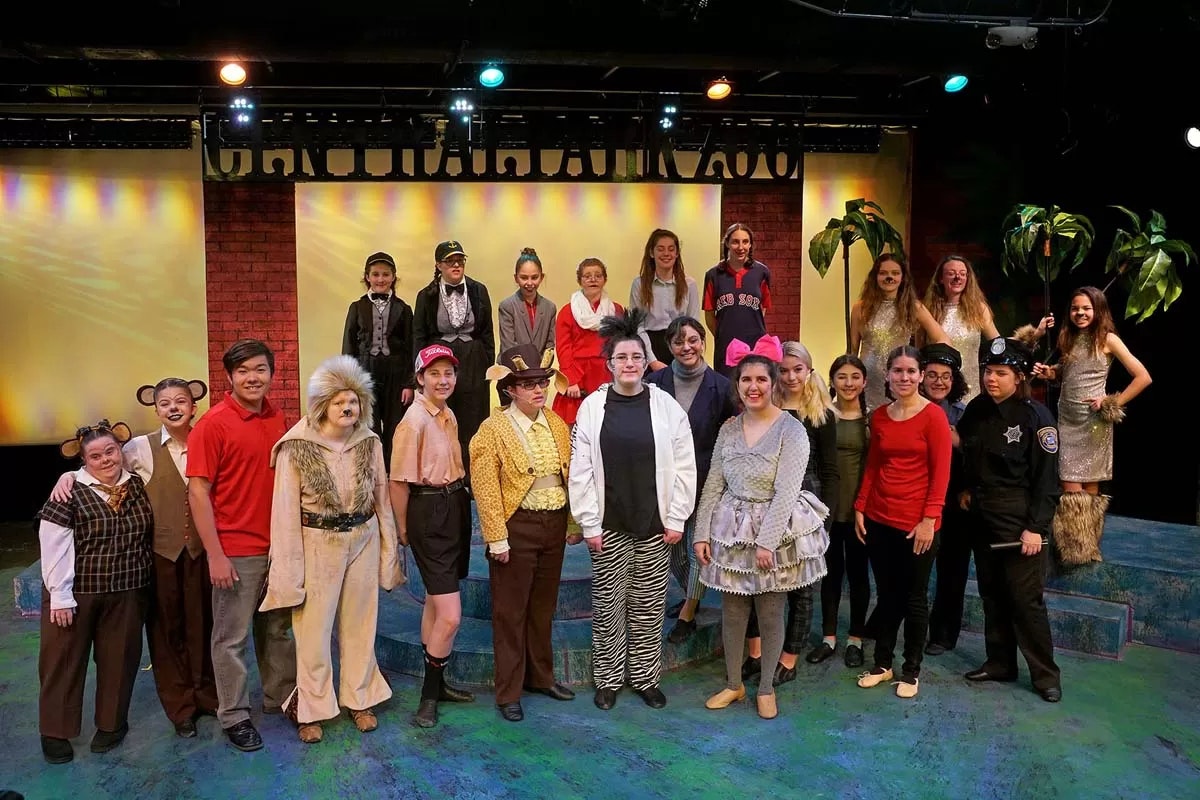 A group of people in costumes pose on a theater stage with a backdrop and stage lighting.