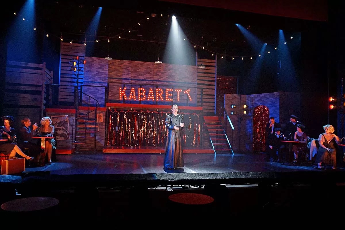 A stage scene from a "Kabarett" show with performers, dramatic lighting, and audience tables.
