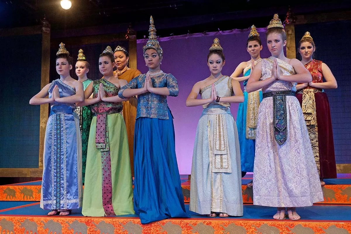 A group of people in traditional attire, wearing ornate headpieces, stand with their palms together in a graceful pose on stage.