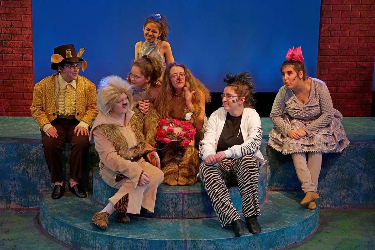 A group of people in colorful, animal-themed costumes sitting on stage steps, some holding flowers, against a blue and red brick backdrop.