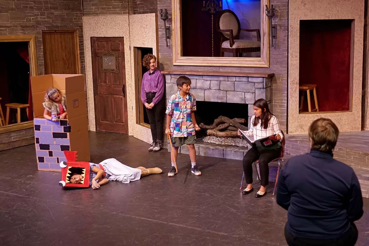 A theatrical play with children performing; one in a dragon costume, another in a cardboard tower, and others acting out a scene on stage.