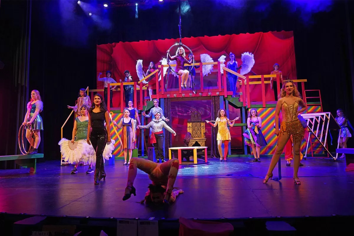 A colorful stage performance with multiple actors in vibrant costumes, a performer suspended on a hoop, and dynamic stage elements.