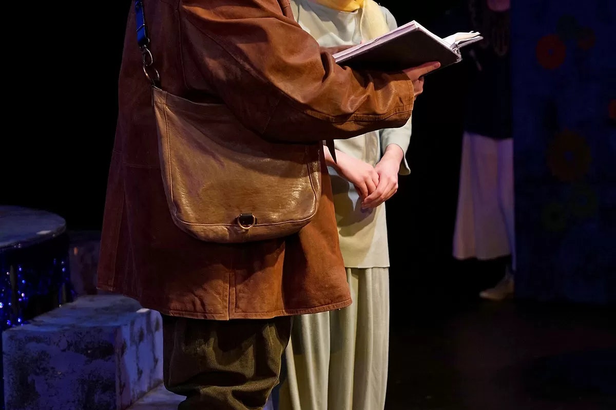 Two people reading a book on stage; one wears a brown jacket and bag, the other in light clothing with hands clasped.