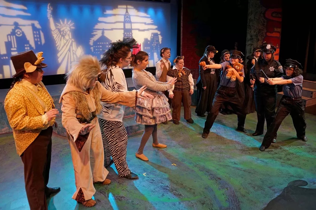 A theater scene with actors in animal costumes and police uniforms, set against a city backdrop with landmarks.