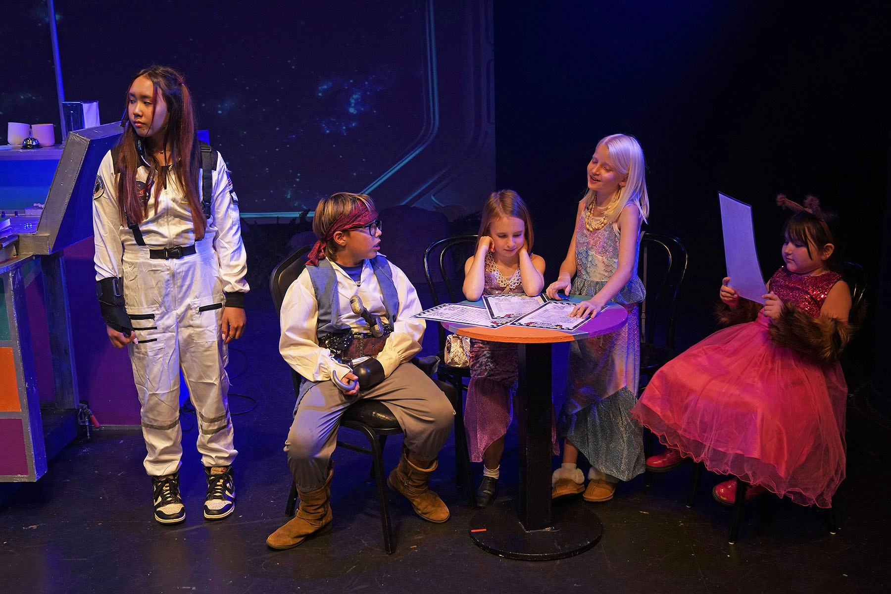 Children in costumes acting on stage: an astronaut, pirate, princesses, and a character with animal ears seated around a table.