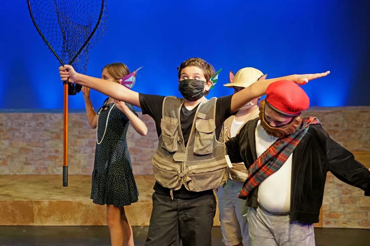 Children performing in costumes onstage; one with a fishing net, others with playful accessories and masks.