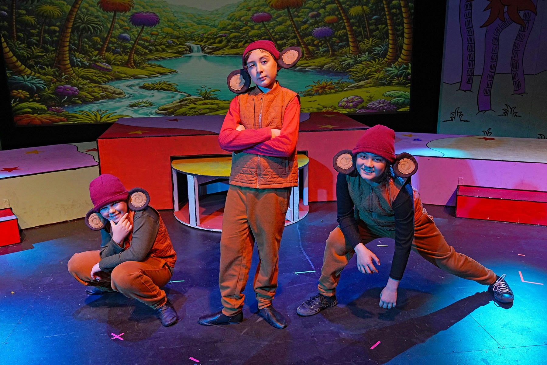 Three children in monkey costumes perform on stage, with a colorful jungle backdrop. They strike playful poses.