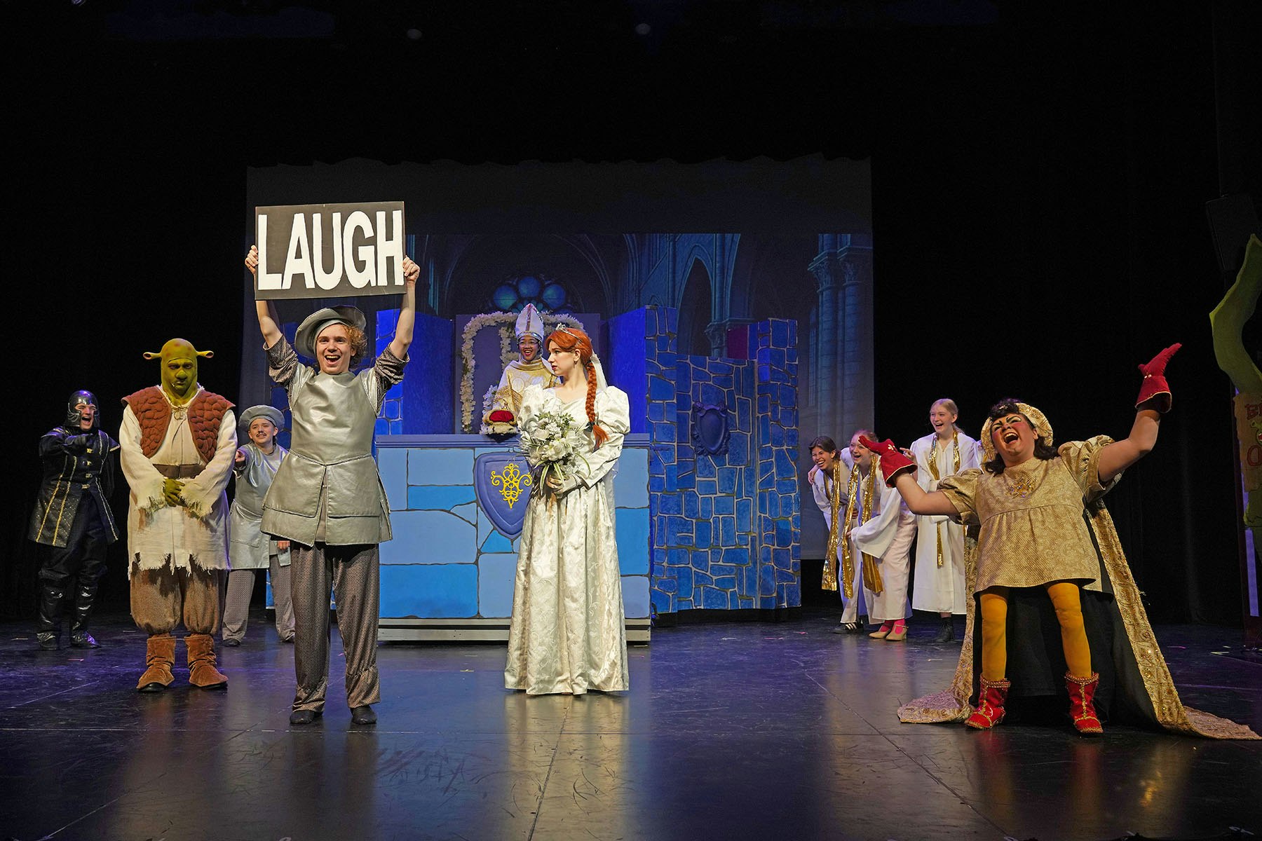 Theater scene with characters in costumes, one holding a "LAUGH" sign, others in medieval-style outfits on stage.
