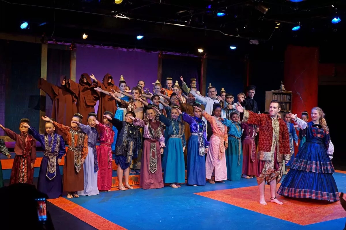 A diverse group of people in colorful costumes performing on stage, many with arms extended outward in unison.