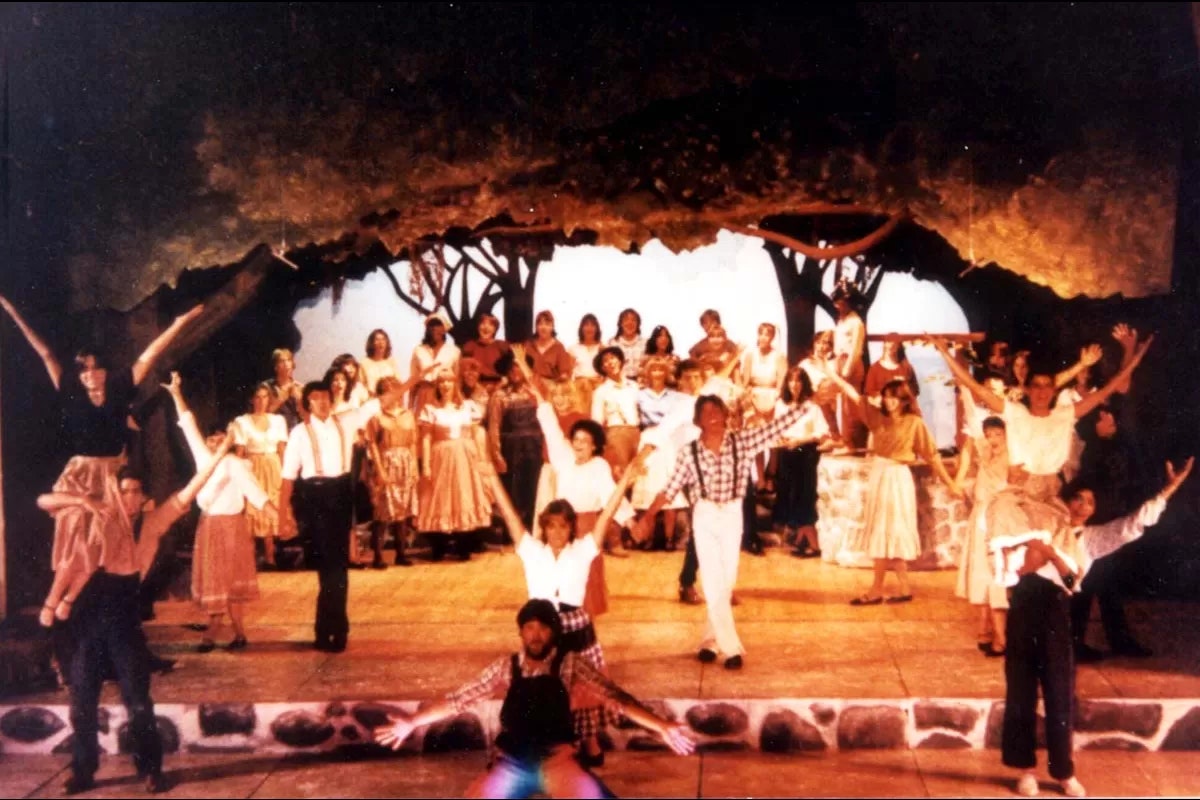 A group of people performing a lively stage musical with colorful costumes and scenic backdrop.