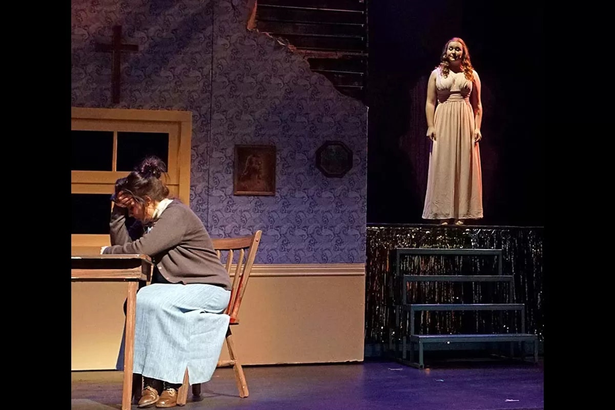 Two people in a theatrical scene: one seated, head in hand, at a table; the other stands on a platform in a long dress, under a spotlight.
