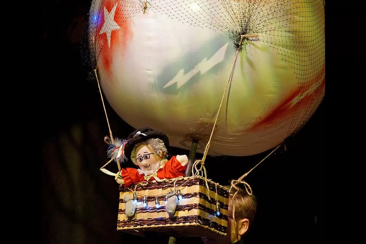 A puppet in a hot air balloon basket, with a large painted balloon above, featuring stars and colors.