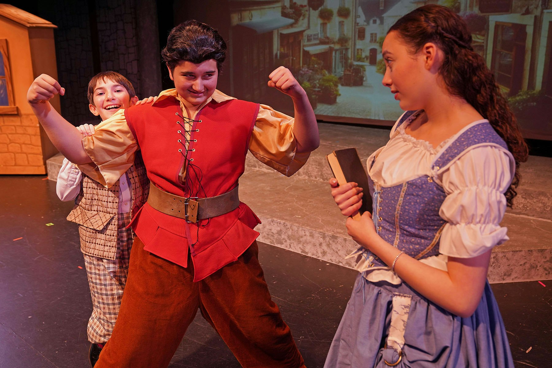 Three people in a theatrical scene, one flexing muscles, another smiling behind, and a third holding a book.