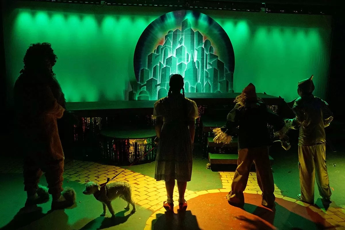Four characters and a dog stand on a yellow brick road, facing an illuminated city backdrop in green light.