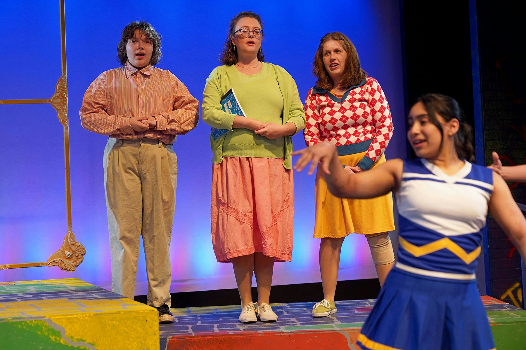 Four people on stage: three in colorful outfits, one in a cheerleader uniform.