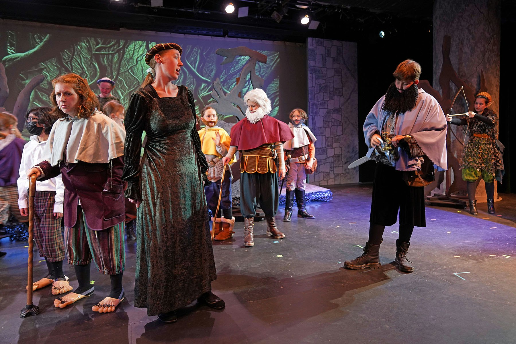 A group of people on stage in a medieval-themed play, wearing costumes like robes, capes, and carrying props like swords and staffs.