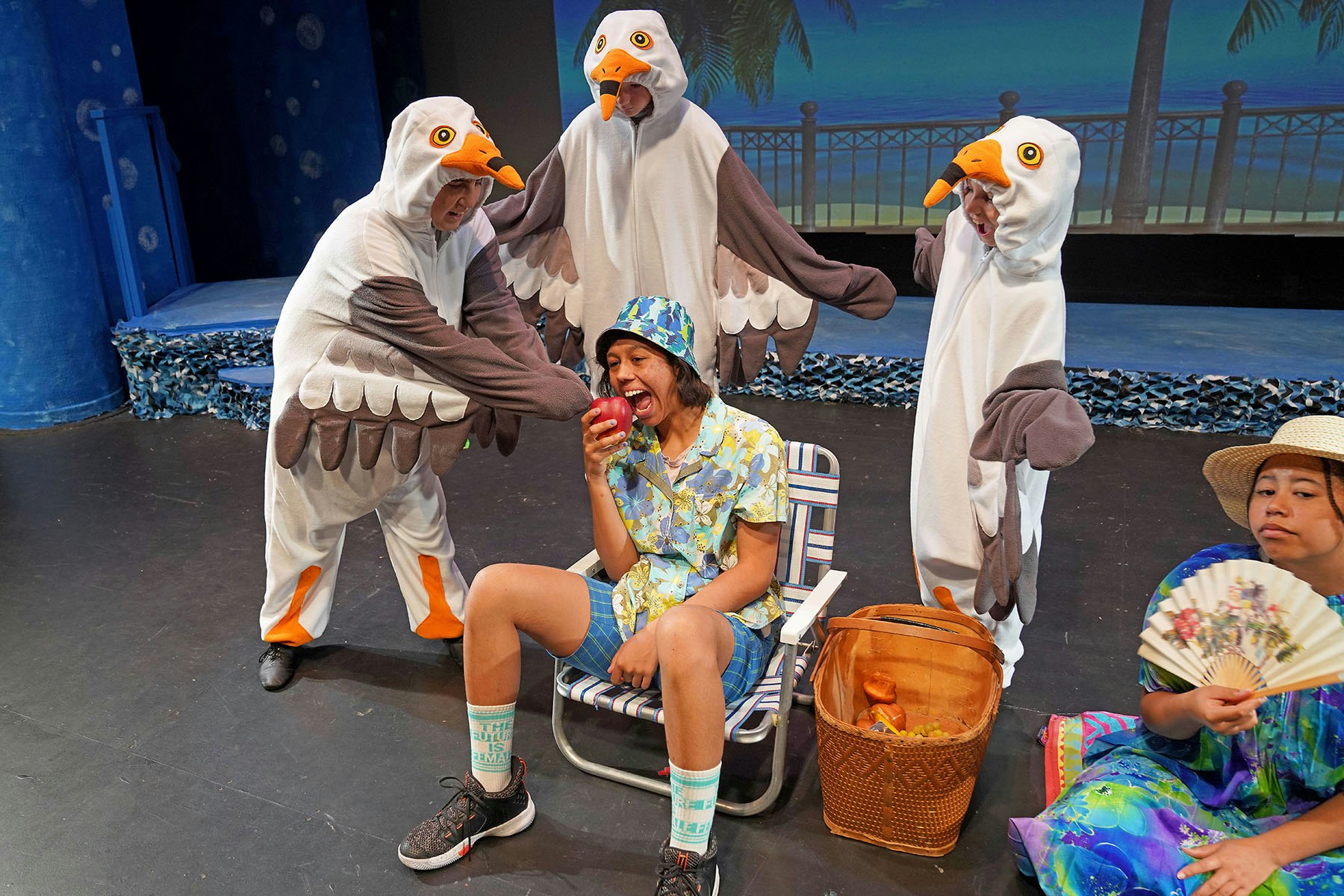 People in seagull costumes surround a person in beach attire eating an apple, with another person holding a fan nearby.