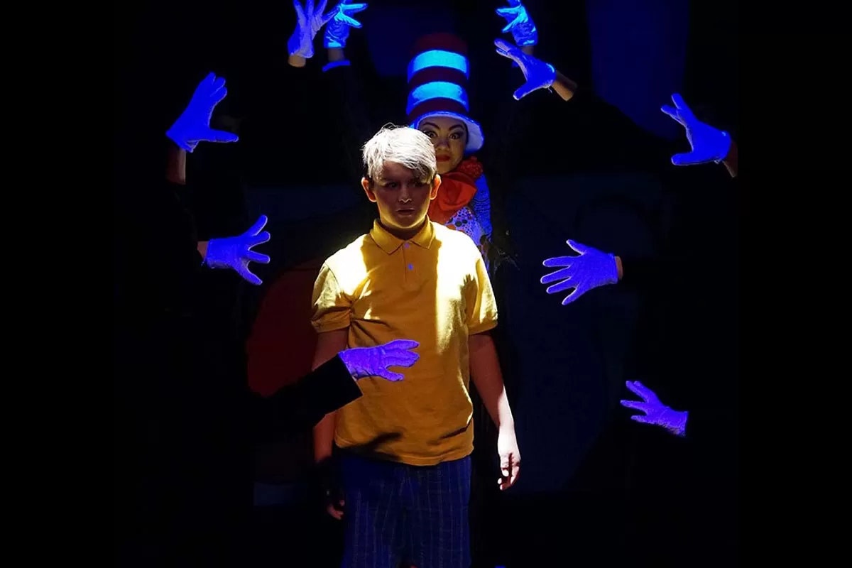 A person in a yellow shirt under spotlight, surrounded by glowing blue gloves, with a figure in a striped hat in the background.