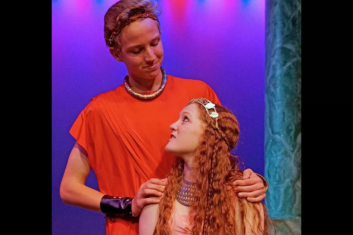 Two people in costume, possibly in a theatrical scene, under colorful stage lighting, looking at each other.
