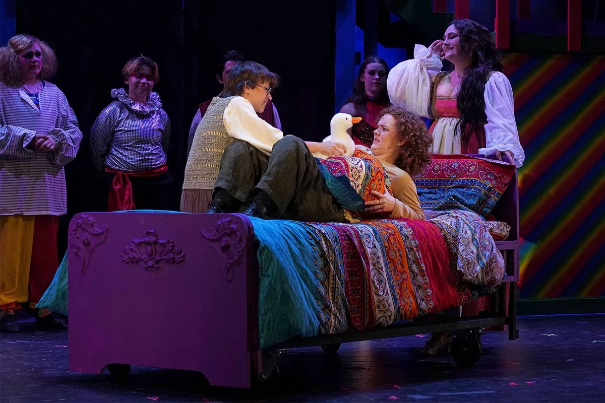 A theatrical scene with people in colorful costumes, one person on a bed, and another holding a puppet or toy animal.