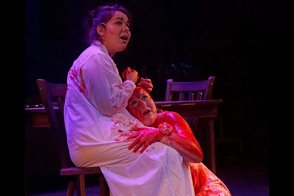 Two people in a dramatic scene, one covered in fake blood, possibly from a stage performance.