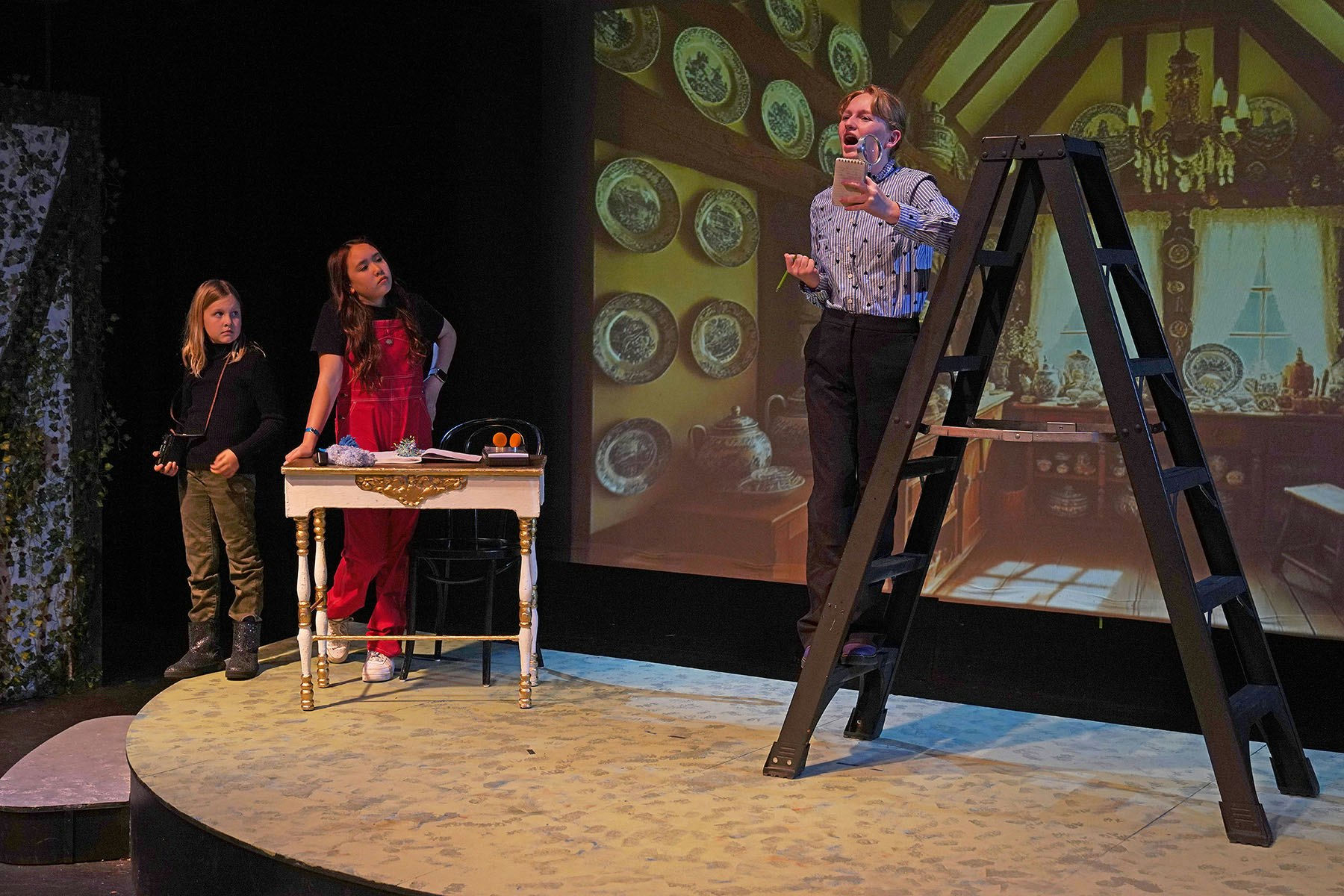 Three people on stage; one stands on a ladder with a notepad, others sit and stand near a table. Background shows a decorated room.