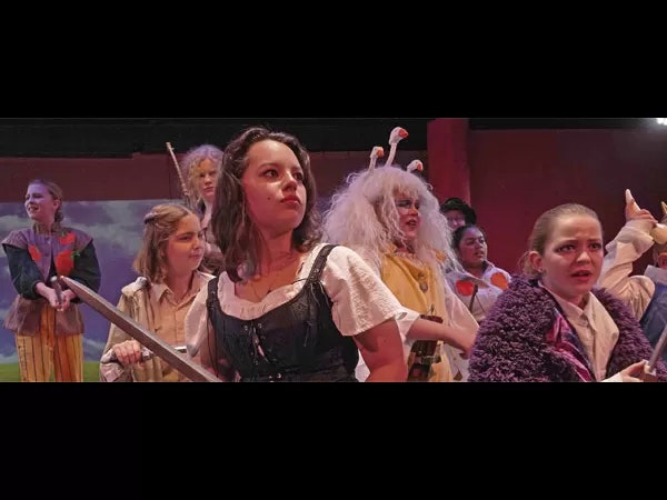 A group of people in theatrical costumes, some holding weapons, are engaged in a dramatic scene on stage.