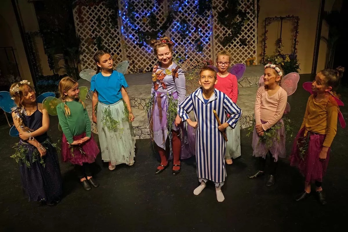 Children dressed as fairies and in pajamas on stage, performing a play with a garden backdrop.