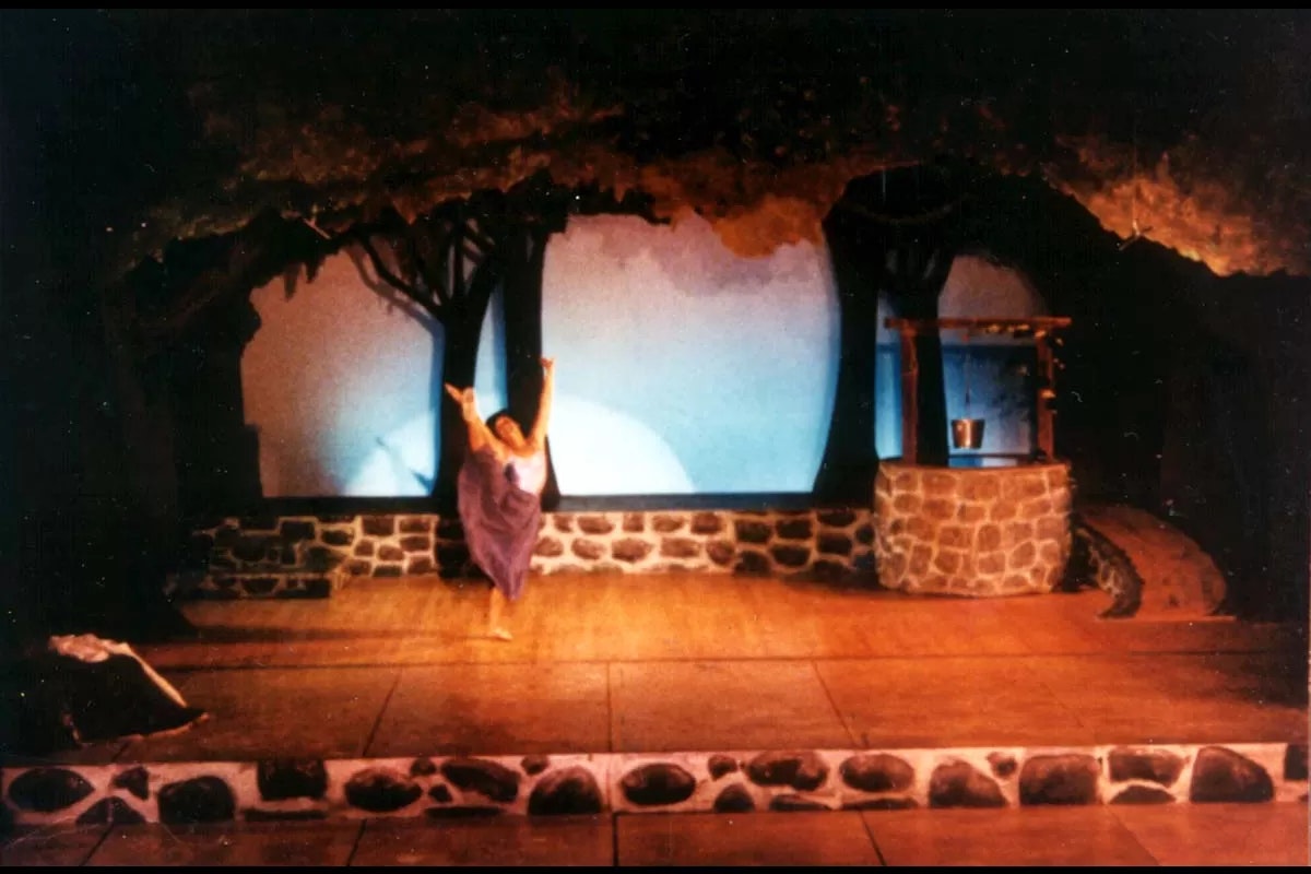 A person in a purple dress dances on a stage set with trees and a stone well backdrop.