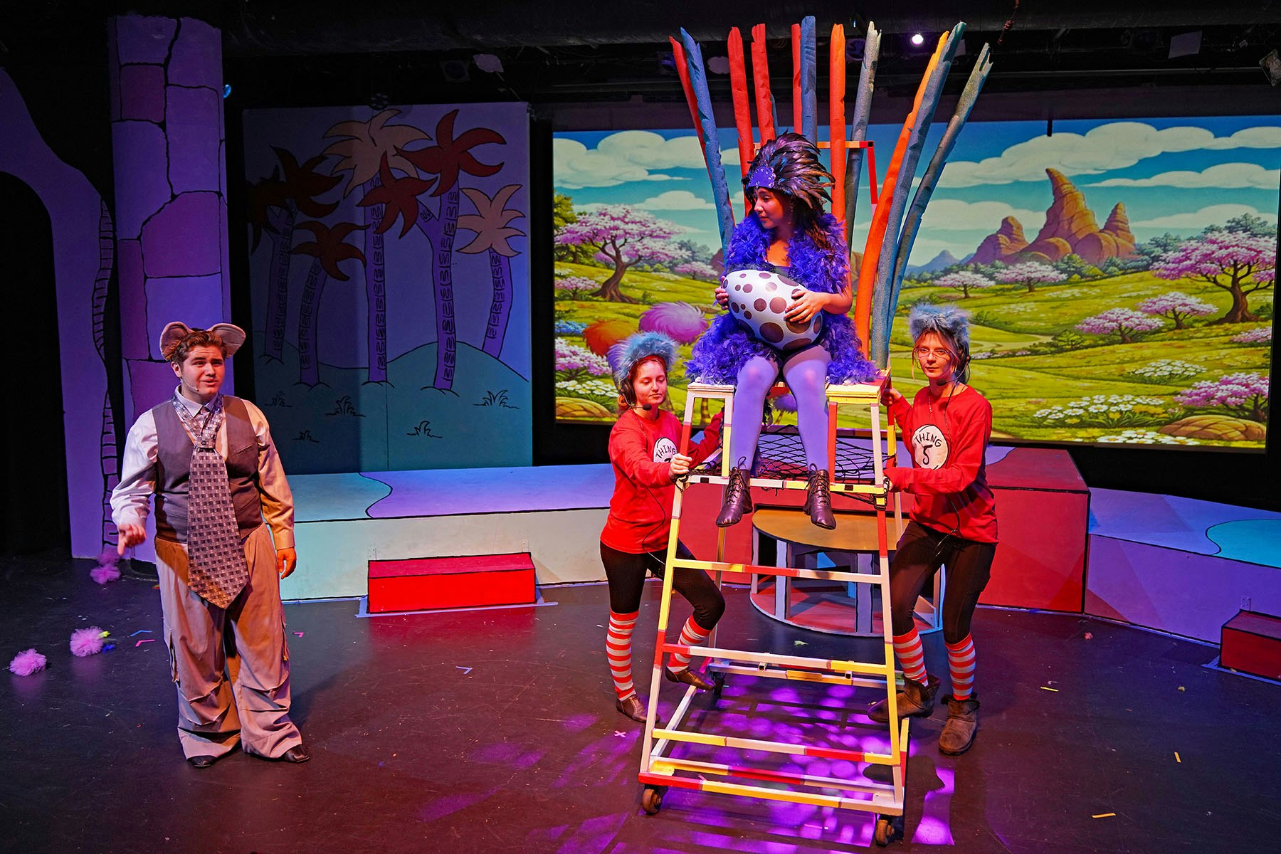 A colorful theater scene featuring performers in costumes, including one on a ladder and a backdrop of whimsical scenery.