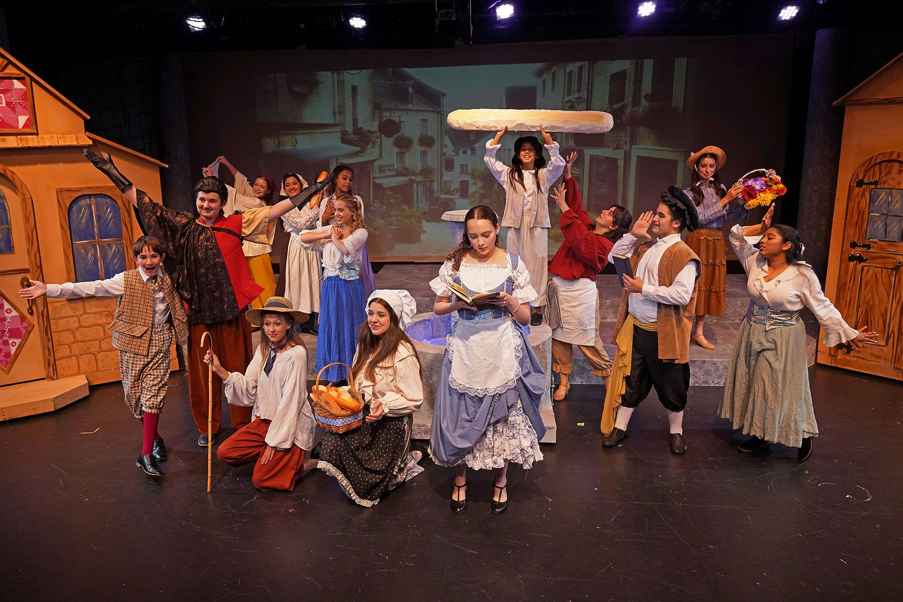 A theater group dressed in period costumes performing on stage with a backdrop of a village scene.