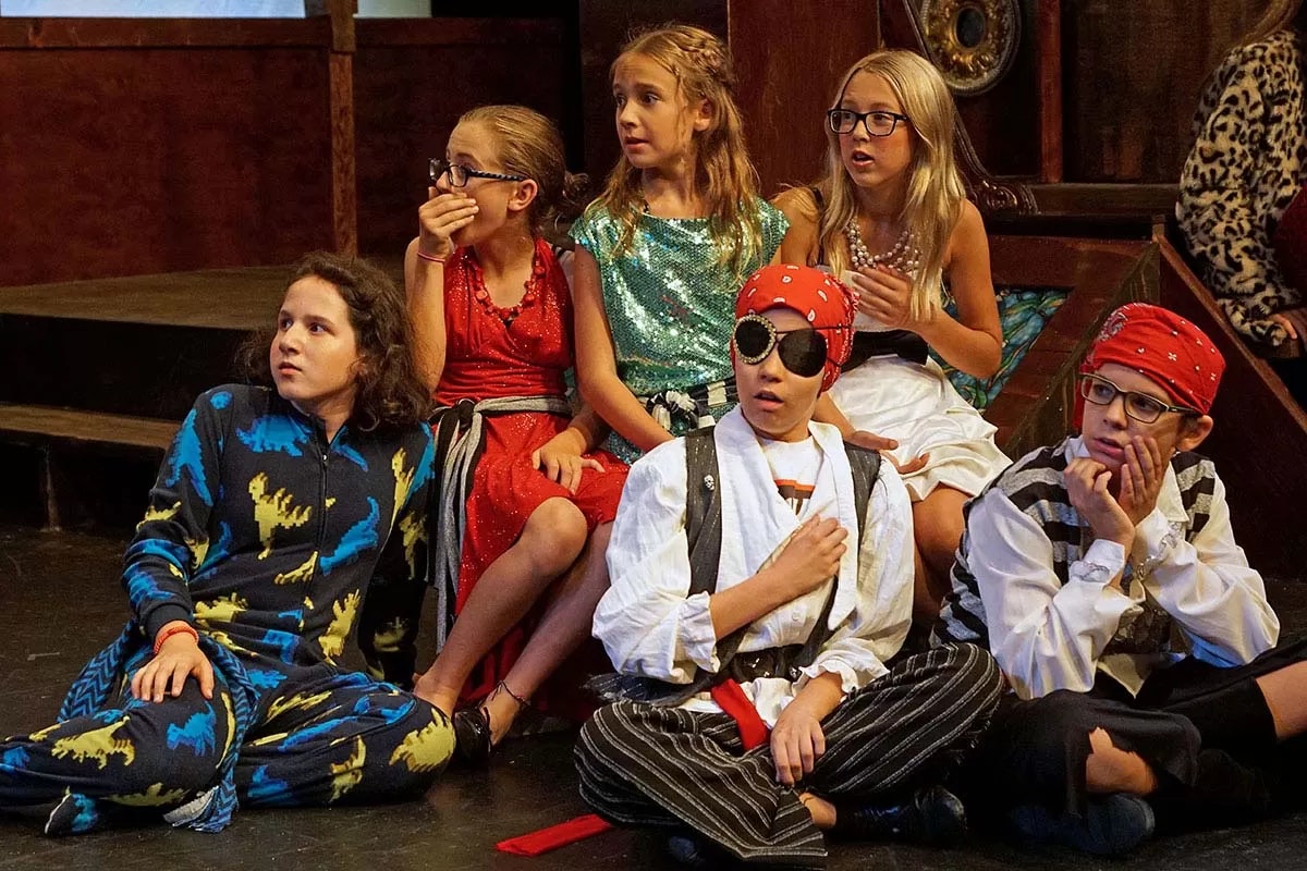 Children dressed in costumes, sitting on stage, looking surprised or curious.