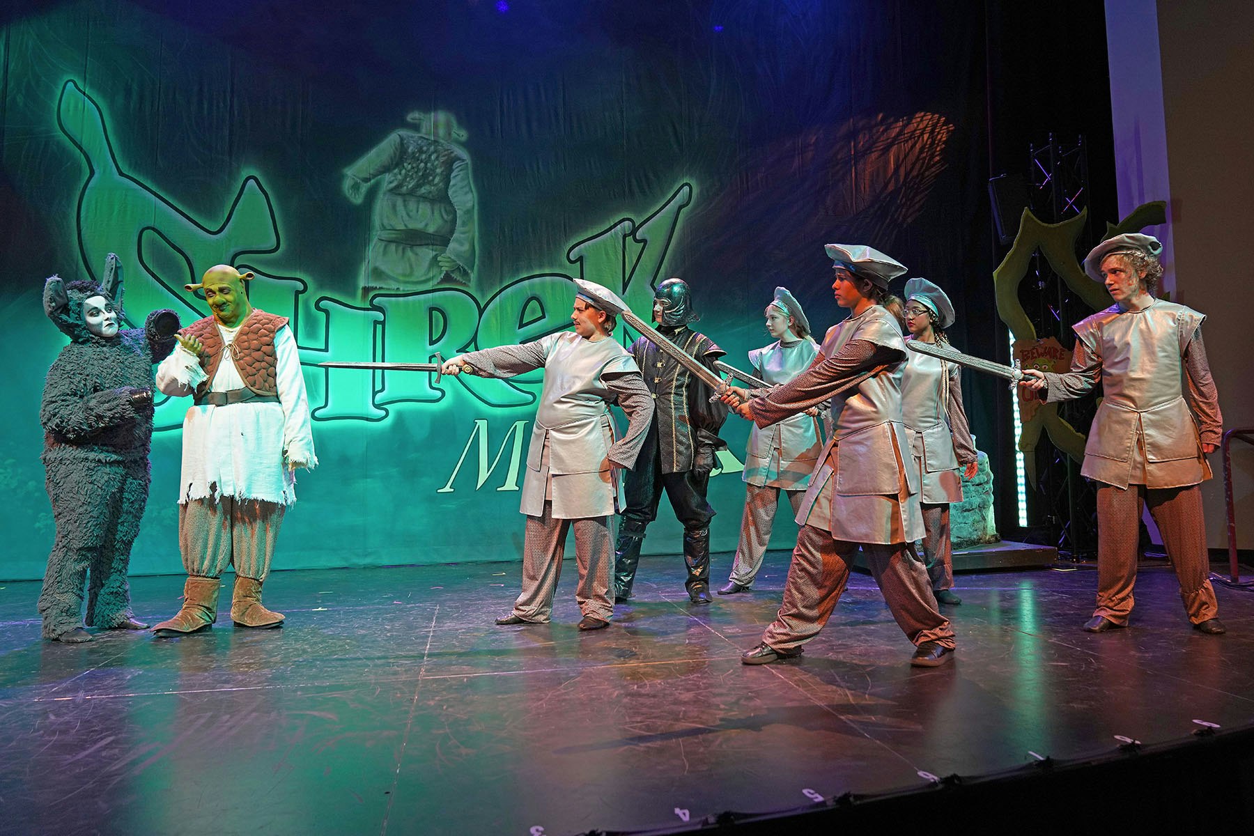 Actors in costumes perform a scene from the musical "Shrek" on stage, featuring Shrek and other characters.