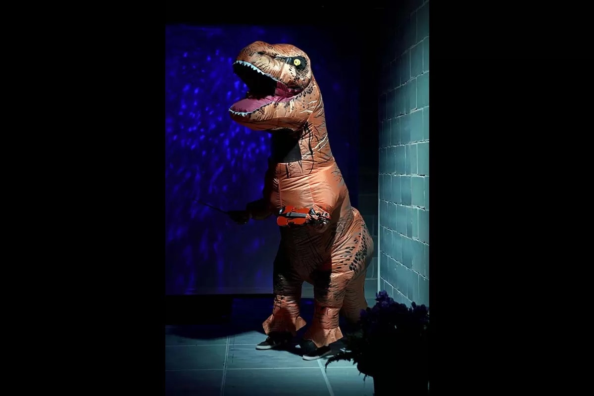 A person in a T-rex costume playing a violin next to a blue-tiled wall.