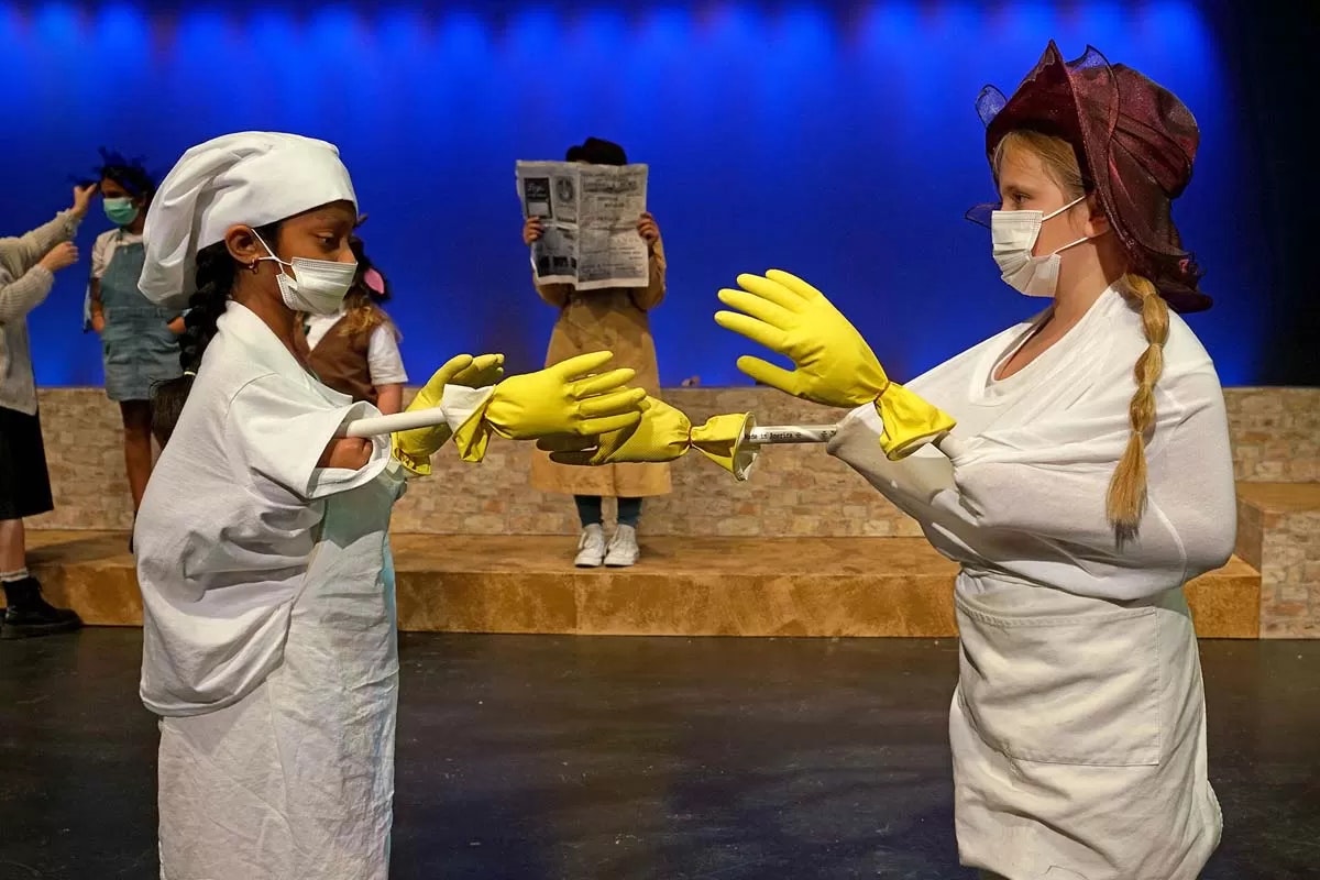 Two people in costume and masks, with oversized yellow gloves, stand face to face on stage. Others are in the background.