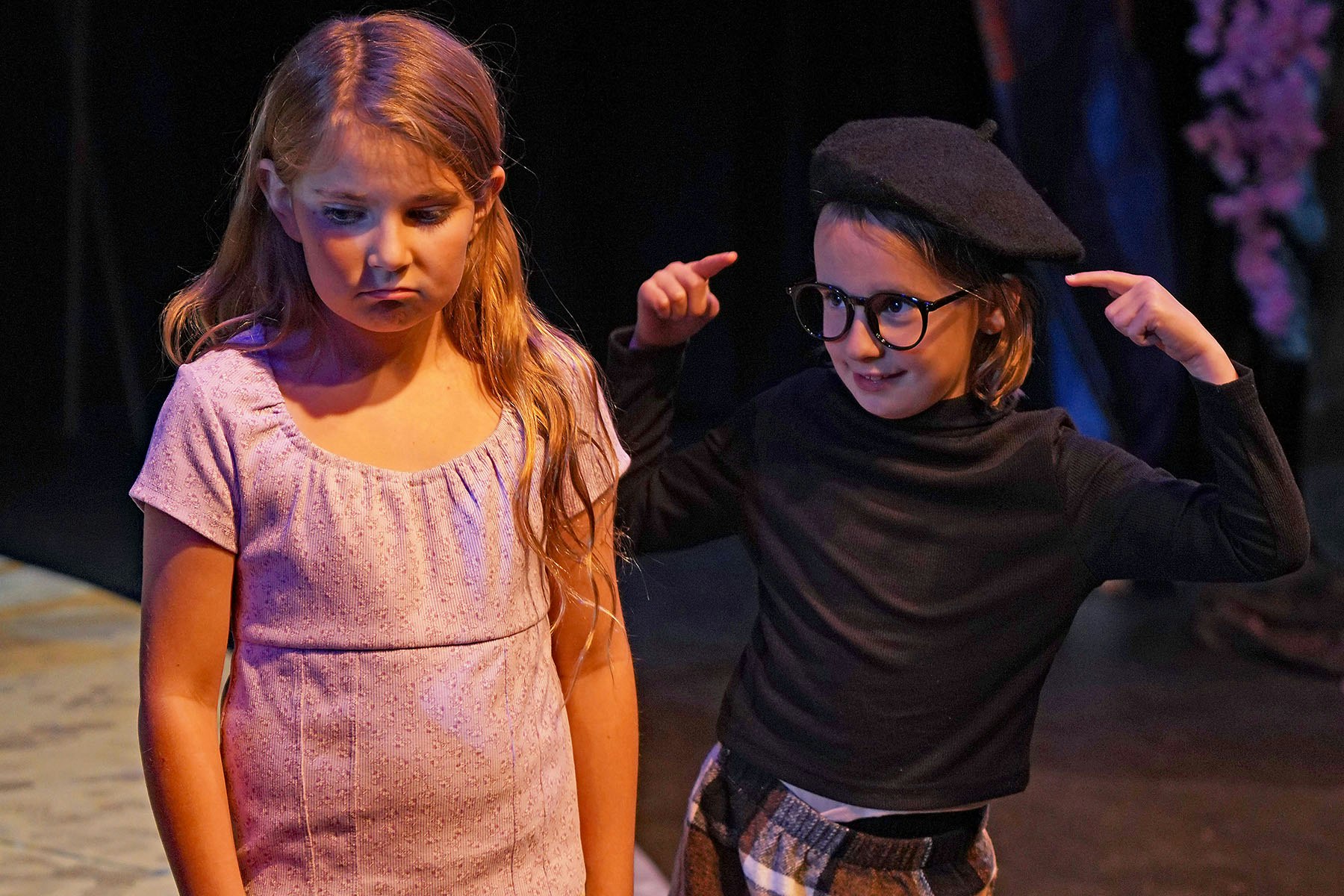 Two children, one looking sad in a dress, the other in glasses and a beret pointing at their head, on a stage.