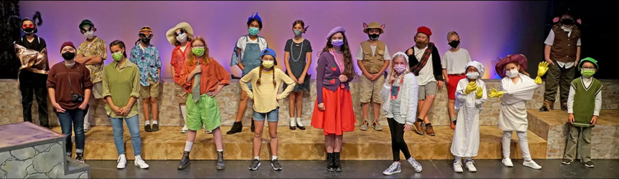 A group of children in colorful costumes and masks stand on stage, posing for a group photo.